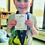 A young child wearing a star medal proudly holds a note indicating that he is a "bucket filler".